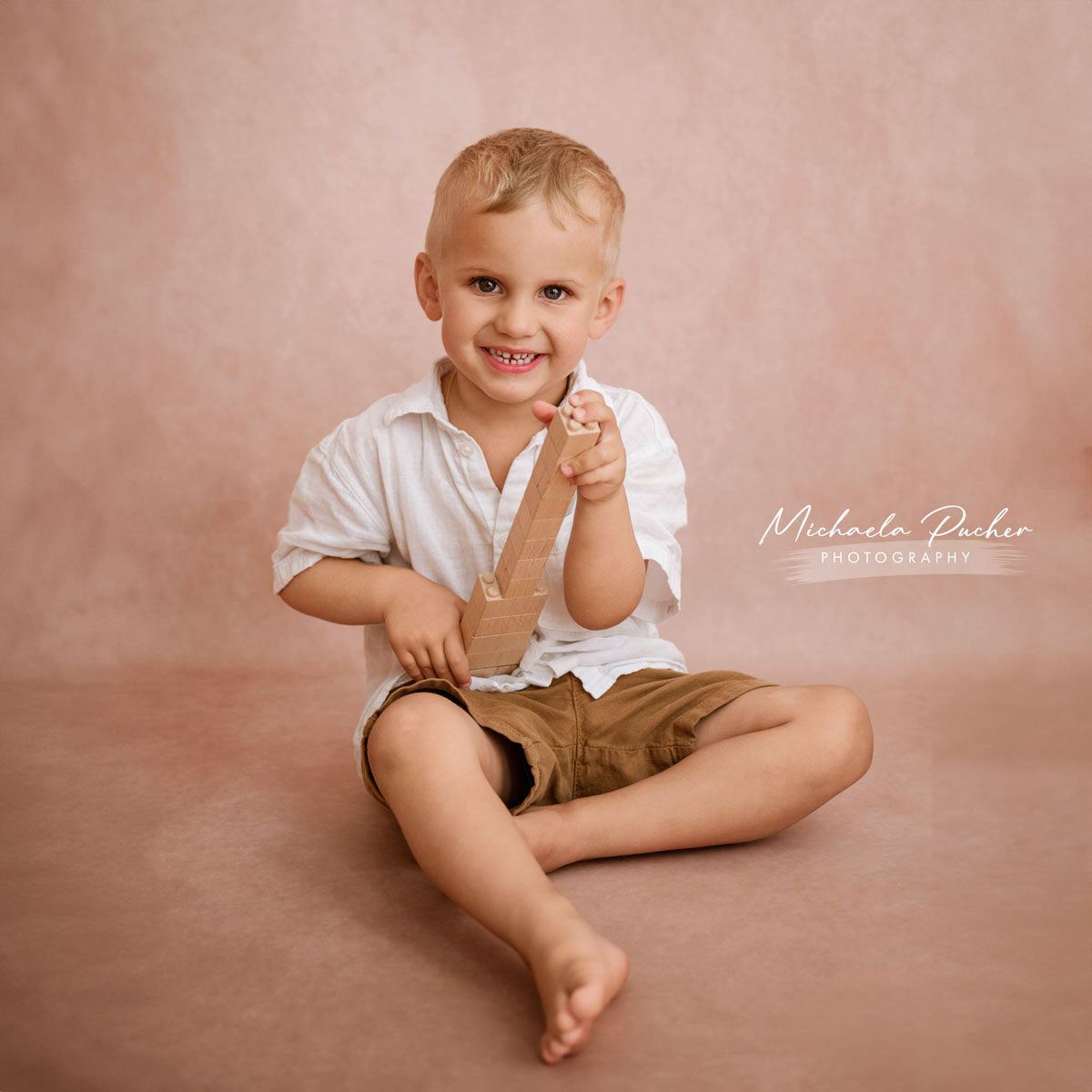 Lachender Junge sitzt barfuß auf dem Boden im Studio und hält ein Holzspielzeug vor einem zarten rosigen Hintergrund.